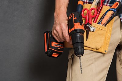 Close up of handyman holding a drill machine with tool belt around waist. Detail of artisan hand holding electric drill with tools isolated over grey background. Closeup hand of bricklayer holding carpentry accessories.
