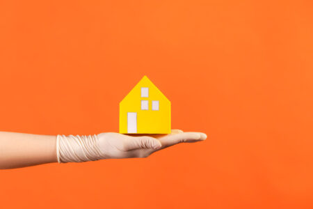profile side view closeup of human hand in white surgical gloves holding yellow paper house exterior in hand.