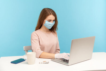 Office worker wearing facial mask and hygienic gloves while typing on laptop, protecting her hands and face to prevent contagious disease, viral infection, coronavirus 2019-ncov. indoor studio shot