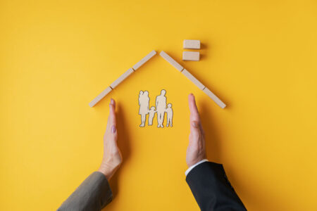 Male and female hands placed to form a home for paper cut silhouette of a family in a conceptual image. Over yellow background.