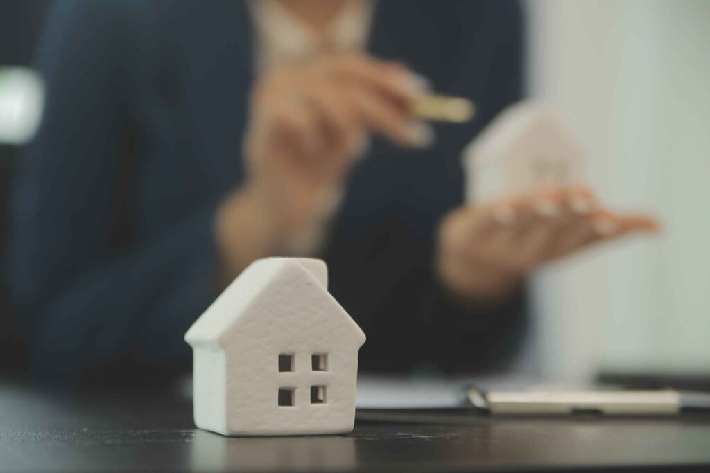 A ceramic model of a house, in the background, blurred out, you can see someone holding a similar model and pointing at it.